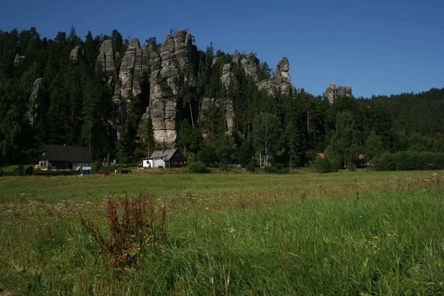 Picturesque landscape with sandstone rocks surrounded by forest, mountain cottages, and a green meadow under a blue sky.