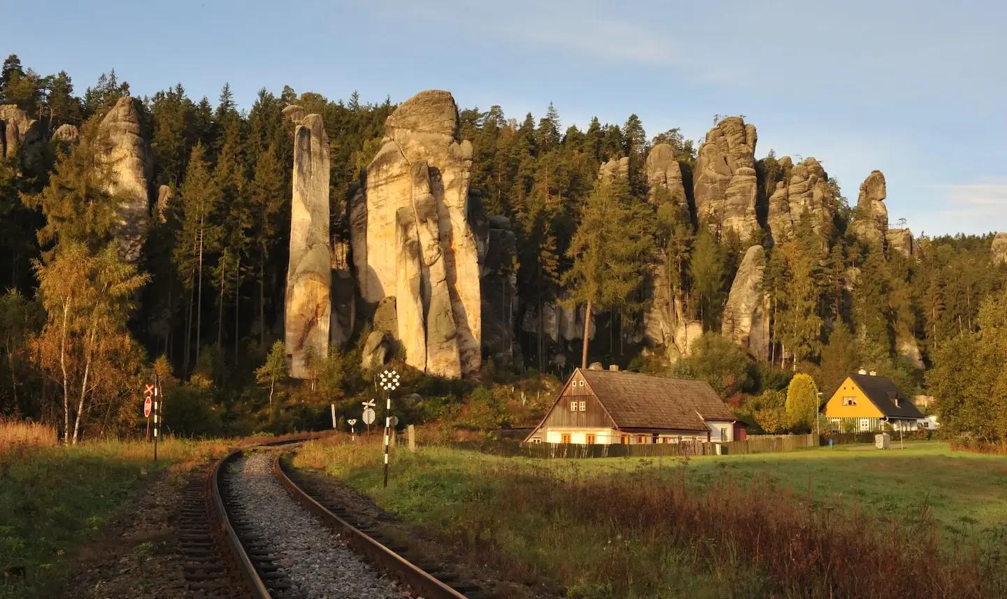 Railway track leading through picturesque landscape between sandstone rocks, forest, and rural houses in warm evening light.